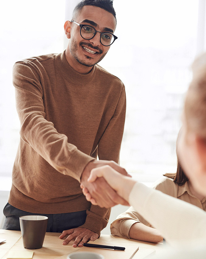 A man with dark hair, glasses and a mustache is seen smiling as he shakes someone's hand who is out of view.